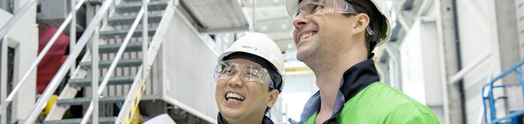 Hymyileviä Valmetin työntekijöitä piirustukset kädessä teollisessa ympäristössä